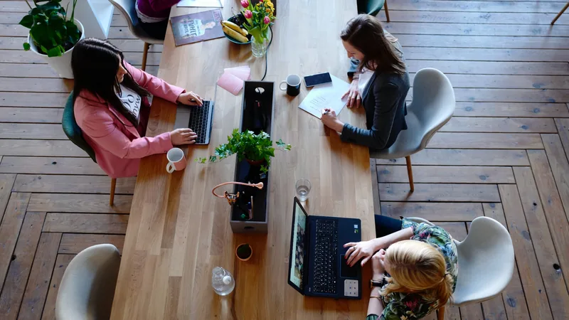 Two women working at laptops across a wooden table, seen from above