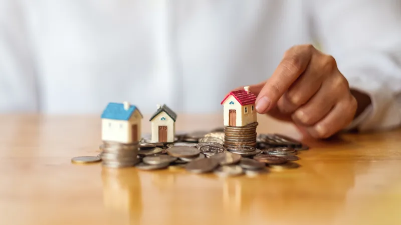 A hand placing a model house on stacks of coins